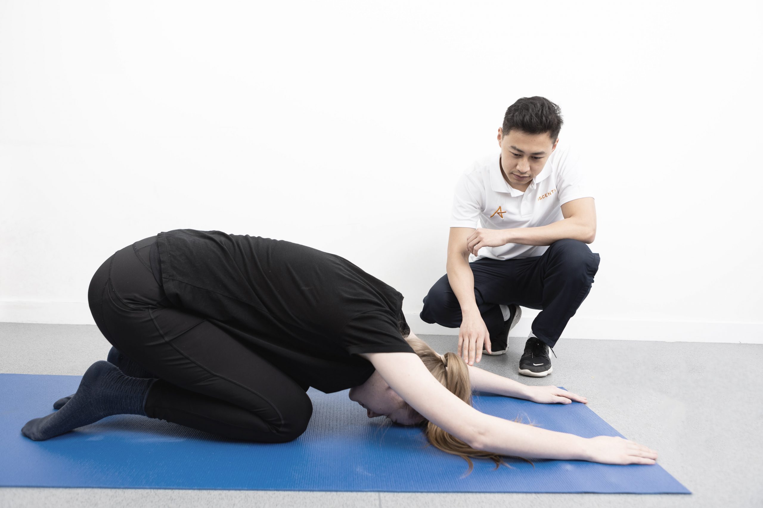 A male physiotherapist dressed in Ascenti uniform (black trousers and white polo shirt) is treating a patient. The patient is dressed all in black, is adopting a stretch pose on a floor mat, her face is not visible.