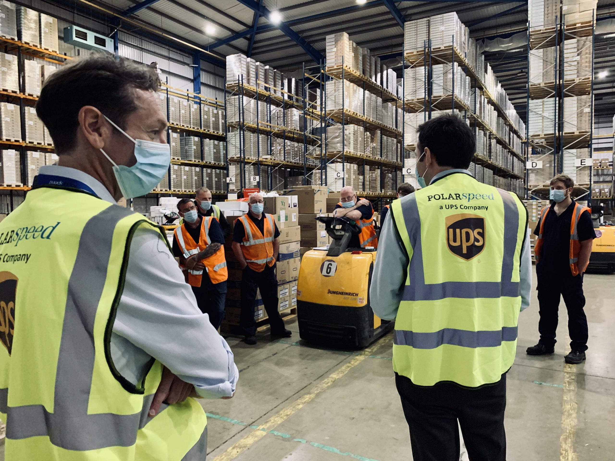 In the foreground we see two men wearing office attire and hi-vis vests, a UPS logo can be seen on the back of the vest. In the background we see a number of warehouse and logistics workers.